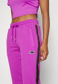 Purple sweatpants with an elastic waistband, featuring side pockets and a black side stripe with embossed lettering, and a small logo accent.