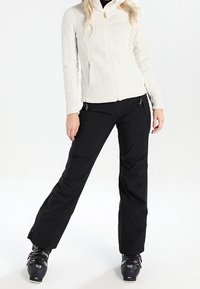 Giacca bianca con texture e zip, abbinata a pantaloni da neve neri. La giacca presenta un colletto alto e una zip dorata. Gli stivali da neve sono neri con dettagli colorati.