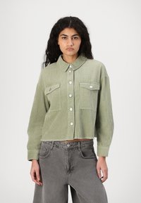 Lys grønn kordfløyelsjakke med krage, sølvknapper, brystlommer og en cropped design. Brukt med grå vidt helse bukser.