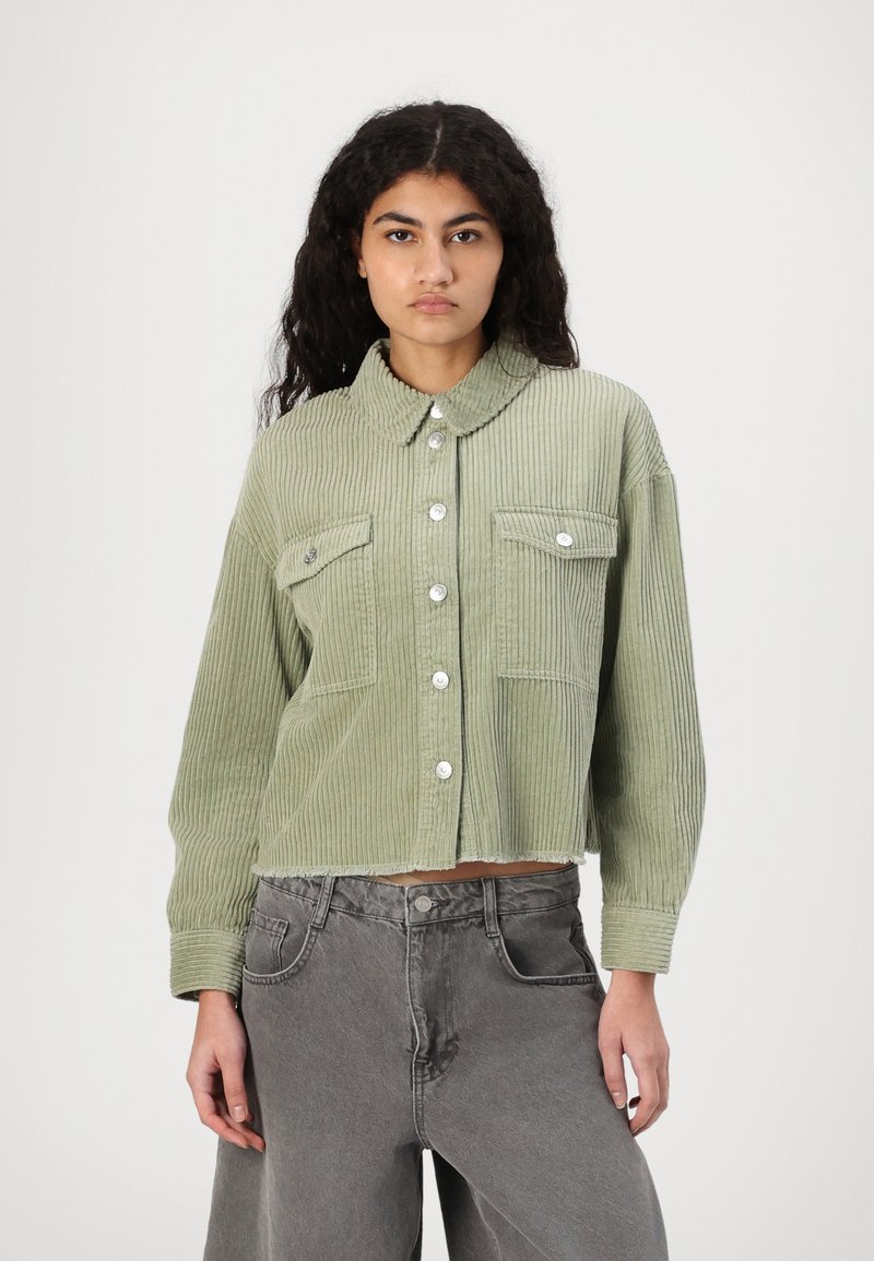 Lys grønn kordfløyelsjakke med krage, sølvknapper, brystlommer og en cropped design. Brukt med grå vidt helse bukser.