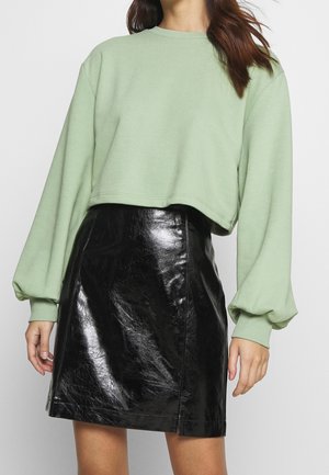 Kobieta ubrana w jasnooliwrowy krótki sweter z bufkami na rękawach i błyszczącą czarną skórzaną mini spódnicę na jednolitym tle.