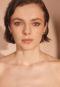 Zlaté kruhové náušnice s červenými príveskami, so štruktúrovaným povrchom. Model má krátke vlasy a neutrálne pozadie. Pleť vyzerá hladko a �čisto.