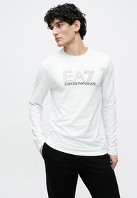 Νέος άνδρας με σγουρά σκούρα μαλλιά, φοράει λευκό πουκάμισο μακρυμάνικο EA7 Emporio Armani και μαύρο παντελόνι, κοιτάζει στο πλάι.