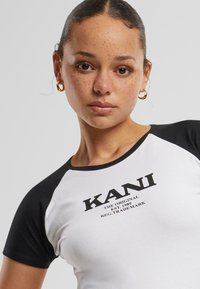 Baltais un melnais īsās piedurknes t-krekls ar apaļu kakla izgriezumu, "KANI" logo tekstu un mīkstu, elastīgu audumu ar pieguļošu formu.
