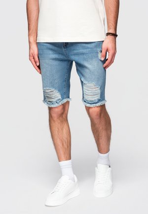 Mand iført lyseblå slidte denimshorts, hvide sokker, hvide sneakers og en hvid skjorte, synlig fra taljen og ned.