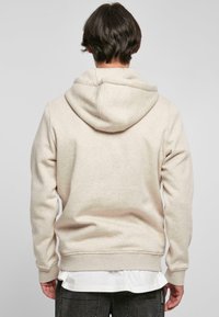 Beige hettegenser med stor justerbar hette, ribbede mansjetter og hem, laget av mykt, teksturert stoff. Båret over en hvit skjorte med lagdelt utseende.