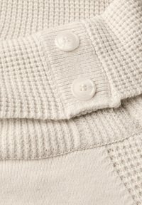 Cremefarbenes Textil mit geripptem und waffelartigem Muster, versehen mit zwei runden weißen Knöpfen, die am Kragenrand kontrastieren.