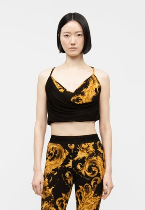 Young woman with short black hair wears black and gold patterned crop top and matching leggings, standing against plain light background.