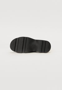 Semelle de chaussure noire avec un motif de bande de roulement rainuré, talon plat, et nom de marque embossé au centre sur un fond neutre.