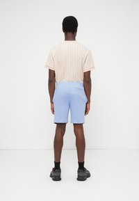 Uomo in piedi di spalle, indossando una camicia beige a maniche corte con motivo, pantaloncini azzurri, calzini neri e scarpe da ginnastica nere su sfondo bianco.