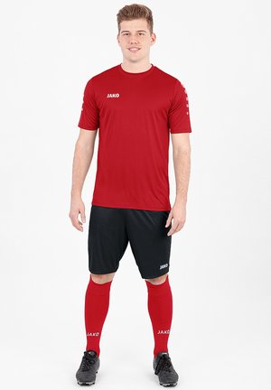 Rotes kurzärmliges Trikot mit Logo, kombiniert mit schwarzen Shorts, roten knielangen Socken und schwarzen Fußballschuhen. Einfaches sportliches Design.