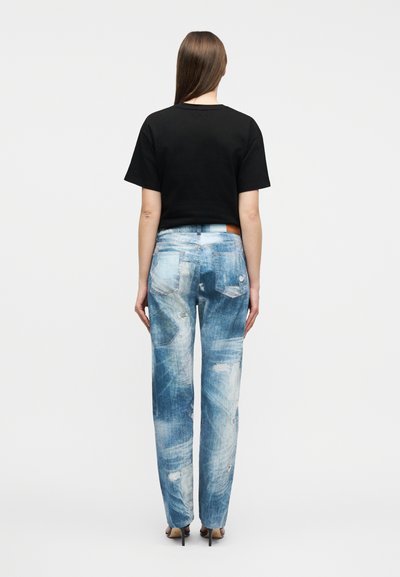 Γυναίκα που στέκεται με την πλάτη προς τα εμπρός, φορώντας μαύρο κοντομάνικο πουκάμισο, ξεθωριασμένο μπλε τζιν με φθορές και μαύρα ψηλοτάκουνα παπούτσια σε λευκό φόντο.