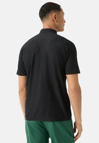 Černé polo tričko s krátkým rukávem a texturovaným povrchem; obsahuje límeček a vertikální design loga na zadní straně. Nosené s zelenými šortkami.