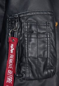 Sektion af sort læderjakke med en lynlåslomme, tekstureret søm og et rødt stofmærke med teksten "REMOVE BEFORE" trykt i hvid.