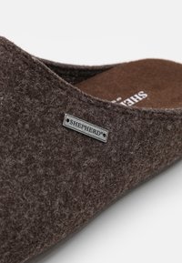 Zapatilla de fieltro marrón con un diseño redondeado, textura suave y una etiqueta de metal marcada "SHEPHERD" en el lateral.