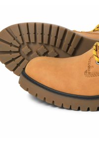 Hnědá kožená bota s drsnou texturou, vybavená silnou gumovou podrážkou s hlubokými vzory pro trakci a žlutými tkaničkami.