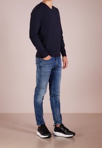 Felpa blu a collo a V, jeans blu e scarpe nere con suole bianche. Il materiale è morbido e la vestibilità è aderente. I dettagli delle cuciture sono visibili.