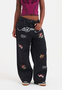 Jeans de mezclilla negra de pierna ancha con varios diseños de parches bordados, que incluyen un logo de marca blanco en la parte trasera y múltiples bolsillos.