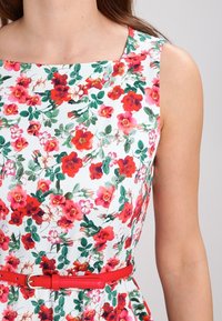 Blumenkleid mit weißem Hintergrund und roten sowie pinken Blumen, einem quadratischen Ausschnitt und einem roten Gürtel, der die Taille betont.