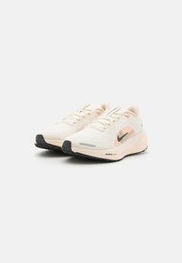 Lehké béžové běžecké boty s síťovinovým svrškem, černým logem Nike, růžovými detaily, texturovanou podrážkou a šněrovacím designem.