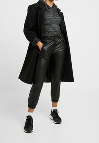 Person i svart lång kappa, grå hoodie, svarta joggerbyxor i konstläder, vita strumpor och svarta sneakers står med händerna i fickorna.