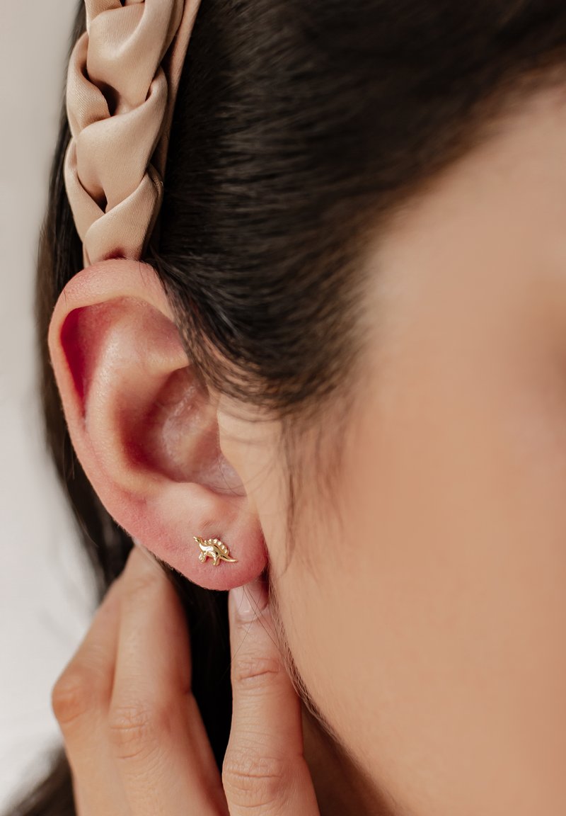 Pendiente de studs en tono dorado con forma de caracol, que presenta detalles texturizados. Se lleva en una oreja perforada, con un accesorio para el cabello de color crema en el pelo.