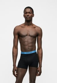 Αθλητικός μαύρος άντρας όρθιος, φοράει μαύρα μπόξερ με μπλε ζώνη που γράφει "BJÖRN BORG", σε απλό ανοιχτό φόντο.