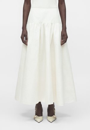 Longue jupe blanche portée par une personne, associée à des chaussures crème pointues avec boucles, debout devant un fond clair uni.