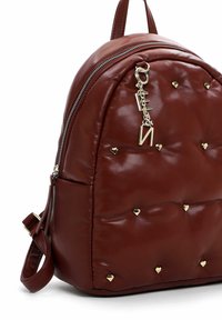 Sac à dos en cuir marron avec des clous en forme de cœur dorés, fermeture éclair, et un porte-clés en forme de lettre dorée suspendu à la tirette supérieure de la fermeture éclair.