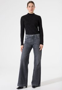 Haut noir à manches longues avec un motif floral texturé, associé à un jean taille haute à jambes larges de couleur gris foncé et des chaussures noires à bout pointu.