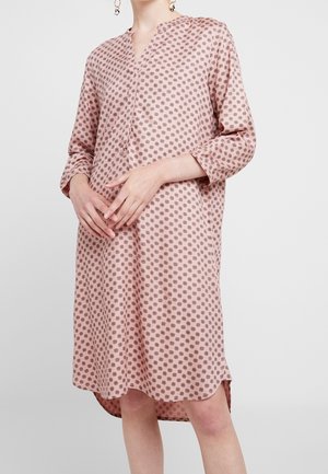 Frau trägt ein locker sitzendes, pinkfarbenes Kleid mit dreiviertel Ärmeln und kleinen dunklen kreisförmigen Mustern, steht vor einem schlichten hellen Hintergrund.