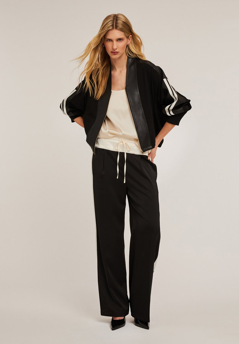 Schwarze Bomberjacke mit Lederfront, weißen Streifen an den Ärmel, kombiniert mit einem cremefarbenen Satinoberteil und schwarzen weit geschnittenen Hosen. Stylische schwarze High Heels.