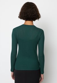 Vihreä ribbineulottu pitkähiha, jossa on vartalonmyötäinen siluetti. Pyöreä pääntie ja teksturoitu kuvio, joka korostaa kevyttä kangasta.