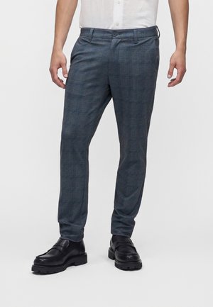 Homme portant un pantalon de costume gris à carreaux slim-fit avec des mocassins en cuir noir et une chemise blanche, debout devant un fond uni.