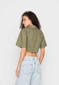 Camisa verde-oliva cortada com colarinho e mangas curtas, feita de um tecido texturizado. Combinada com jeans de cintura alta azul clara.