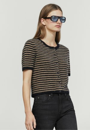 Femme portant des lunettes de soleil teintées bleues, un cardigan rayé marron et noir à manches courtes boutonné, et un jean noir taille haute sur un fond uni.