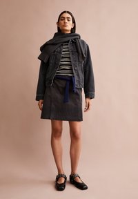 Donker denimjacket over een zwart-wit gestreepte trui, gecombineerd met een grijze rok met een blauwe riem. Zwarte schoenen met metalen accenten.