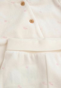 Miękki, kremowy strój dla niemowląt z delikatnymi różowymi wzorami i drewnianymi guzikami na górze, połączony z pasującymi spodniami na elastycznej talii.