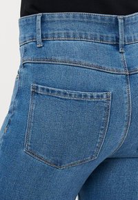 Vaqueros de mezclilla azul de talle alto con una textura suave, bolsillo trasero y detalles de costura visibles a lo largo de la cinturilla y las costuras.