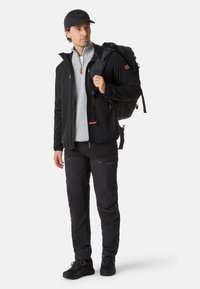 Man wearing black cap, black jacket, gray sweater, black pants, and black hiking shoes carrying a large black backpack, looking right.