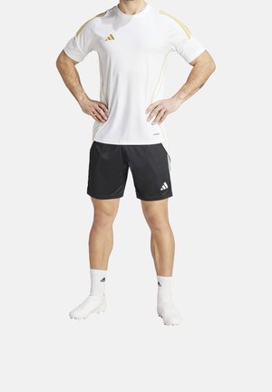 Mężczyzna sportowiec stojący z rękami na biodrach, ubrany w białą i złotą koszulkę z krótkim rękawem, czarne spodenki, białe skarpetki oraz białe korki.