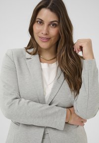 Blazer léger gris clair avec des revers structurés, en tissu doux et à manches longues, associé à un top blanc texturé et un collier en chaîne dorée.