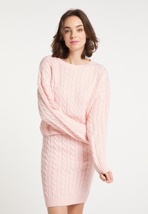 Young woman with brown hair wearing a light pink cable knit sweater and matching skirt stands against a white background.