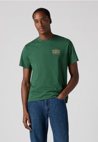 Groene katoenen t-shirt met korte mouwen en een ronde hals. Bevat gele geborduurde tekst op de borst. Geselecteerd in combinatie met blauwe denim jeans.