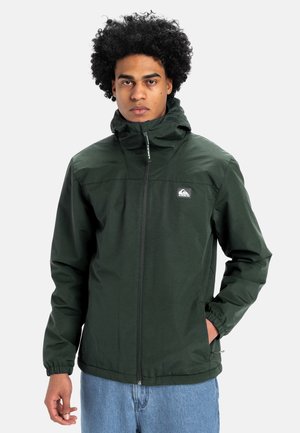 Joven con chaqueta con capucha verde oscuro con cremallera frontal y vaqueros azul claro, de pie contra un fondo claro y liso.