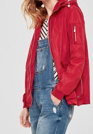 Kobieta ubrana w czerwoną kurtkę na zamek, nałożoną na koszulę w jodełkę i niebieskie dżinsowe ogrodniczki, z ręką w kieszeni, na jednolitym tle.