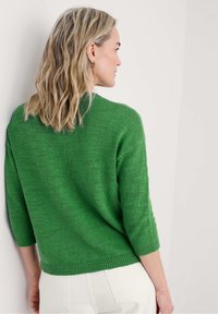 Zaļa adīta svārku topiņš ar apaļu kakla izgriezumu, 3/4 garuma piedurknēm un ribota apmale. Gluda tekstūra ar maigu rakstu, apvienota ar baltām biksēm.