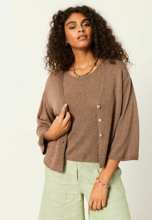 Woman with curly hair wearing a brown knit cardigan over a matching top and light green pants, accessorized with a necklace and bracelet.
