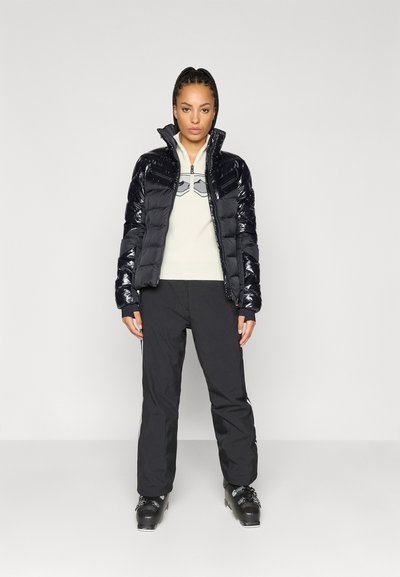 Μαύρο γυαλιστερό μπουφάν με υψηλό γιακά, συνδυασμένο με ένα ανοιχτόχρωμο φλις και μαύρο σκι παντελόνι. Μαύρες μπότες σκι ολοκληρώνουν την εμφάνιση.