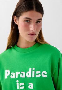 Felpa verde con la scritta "Paradise is a" in bianco, con collo rotondo e polsini a costine, realizzata in un morbido tessuto texturizzato.